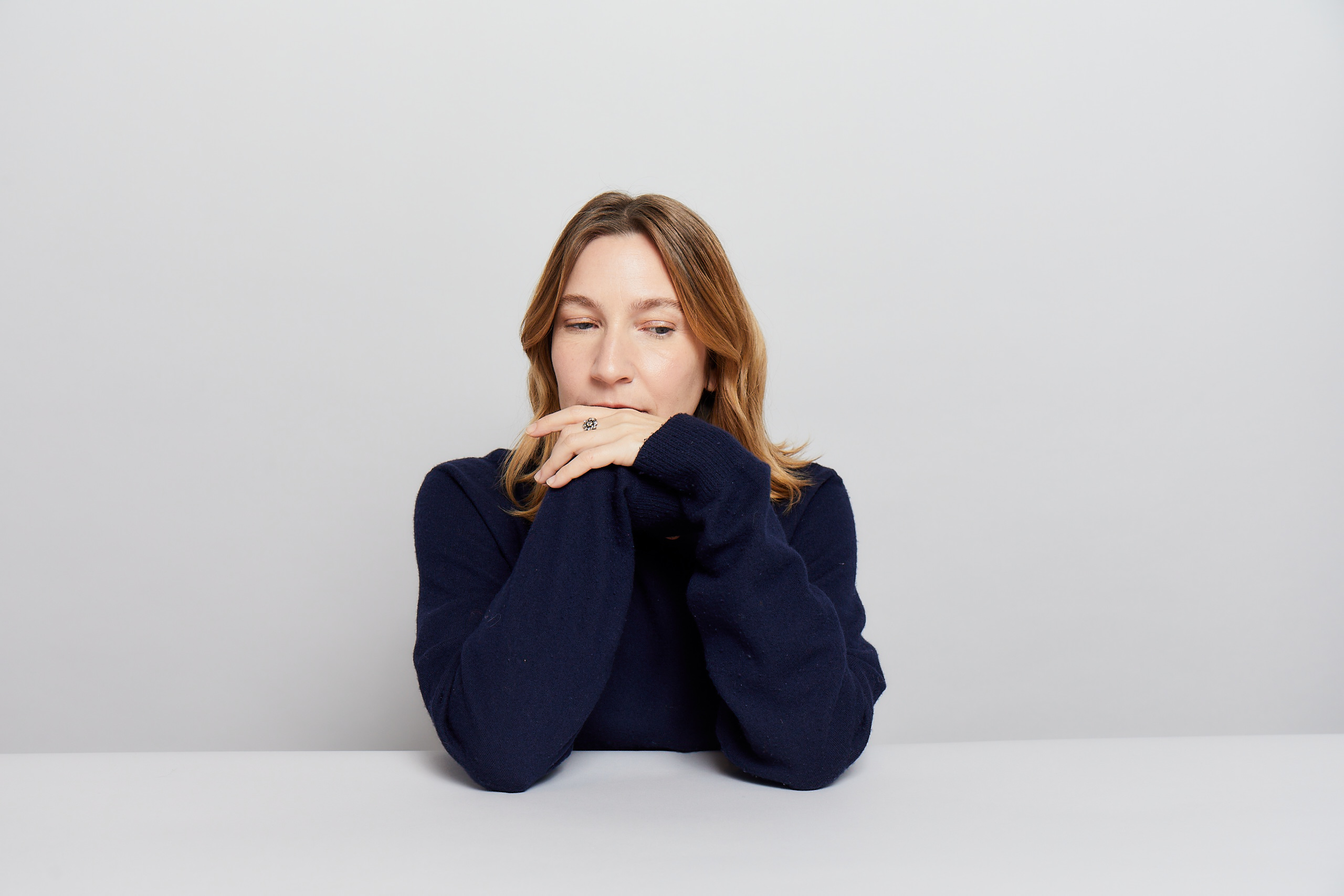 Woman with long hair rests her chin on crossed hands, wearing a dark sweater, sitting at a white table against a plain white background. She appears thoughtful, gazing slightly downward.