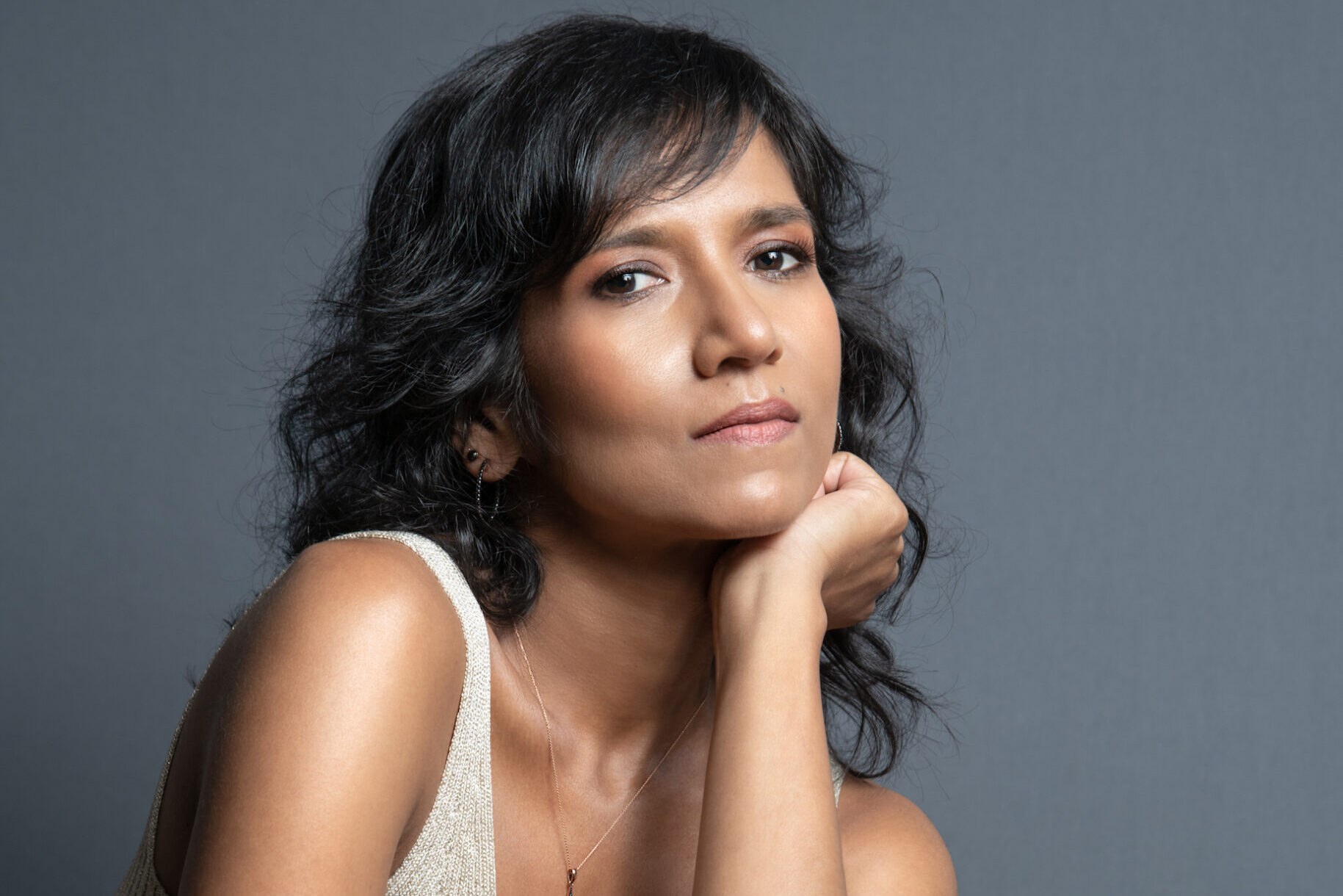 A woman with wavy black hair sits against a plain gray background. She rests her chin on her hand, wearing a sleeveless beige top and black pants, with a star-shaped necklace. Her expression is contemplative.