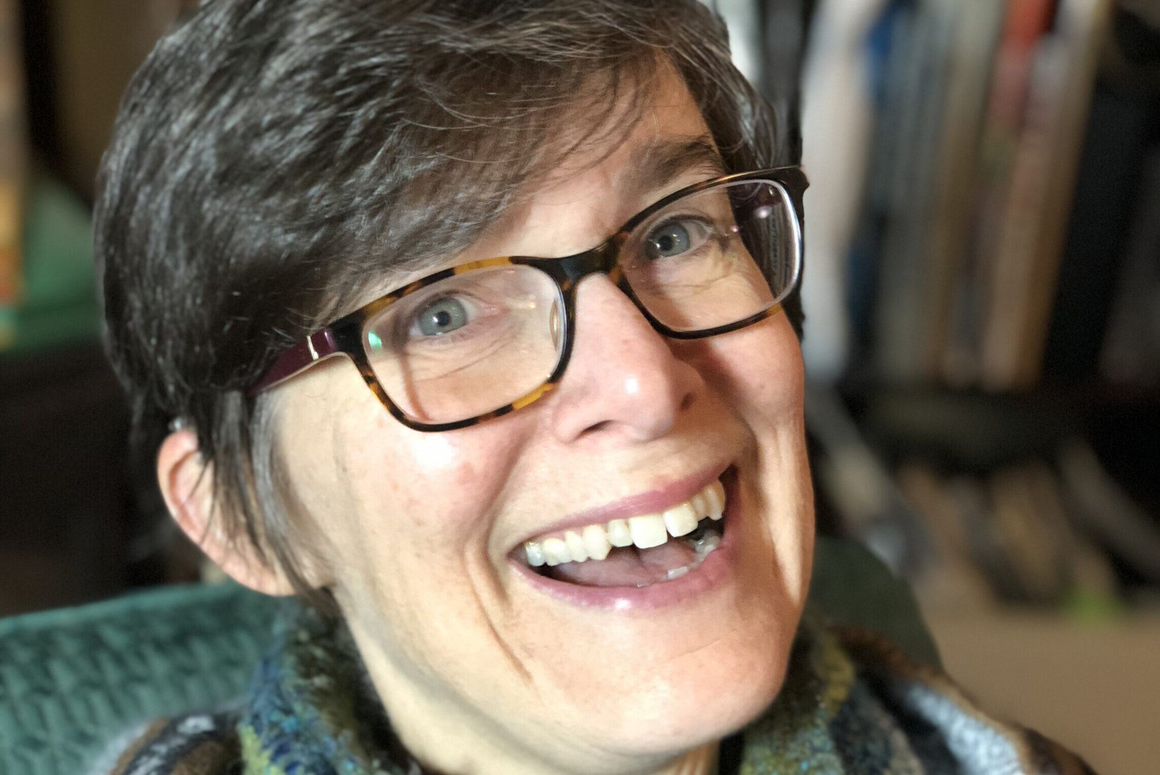 A person with short hair and glasses smiles warmly at the camera. They are wearing a cozy striped sweater and a multicolored scarf, sitting indoors with a bookshelf in the background.