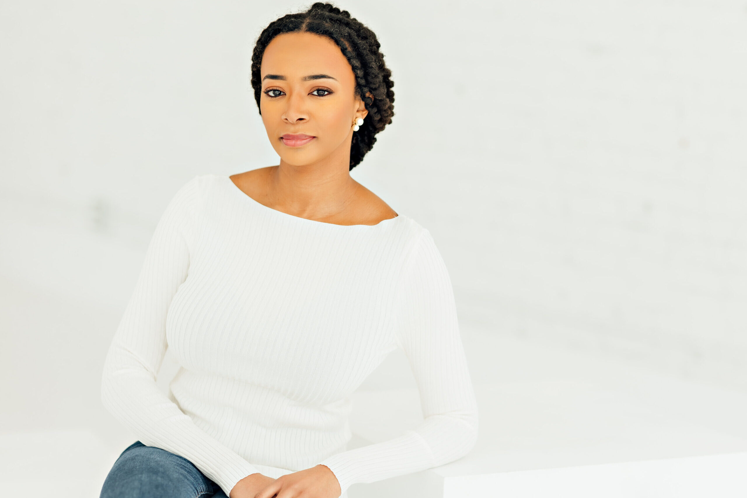 Woman with braided hair, wearing a white sweater and jeans, sitting on a white surface against a minimalistic, light background. She has a calm expression and her hands are gently clasped.