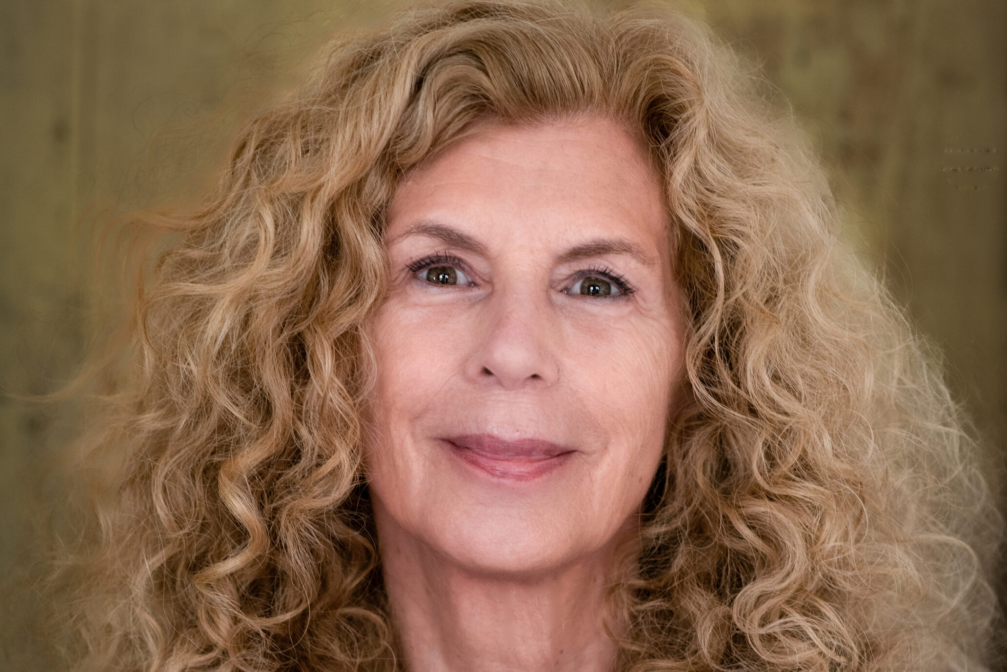 A person with long curly blond hair smiles slightly while looking at the camera. They wear a red top and a gold necklace, set against a blurred brownish background.