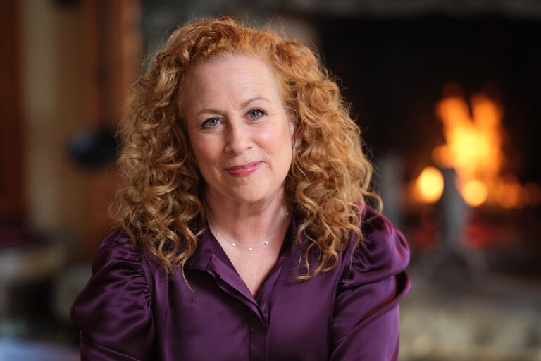A person with curly red hair sits on a brown leather chair, wearing a purple blouse and black pants. There’s a lit fireplace in the background, creating a warm and cozy atmosphere.