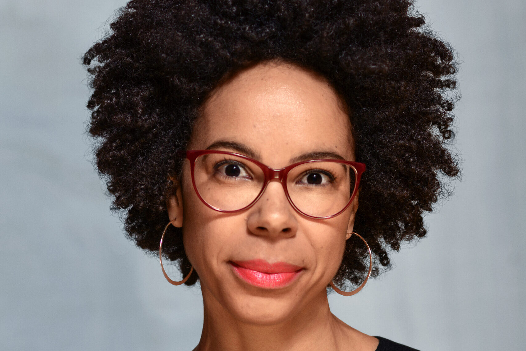 A person with curly hair and red glasses wearing a black top, against a plain background.