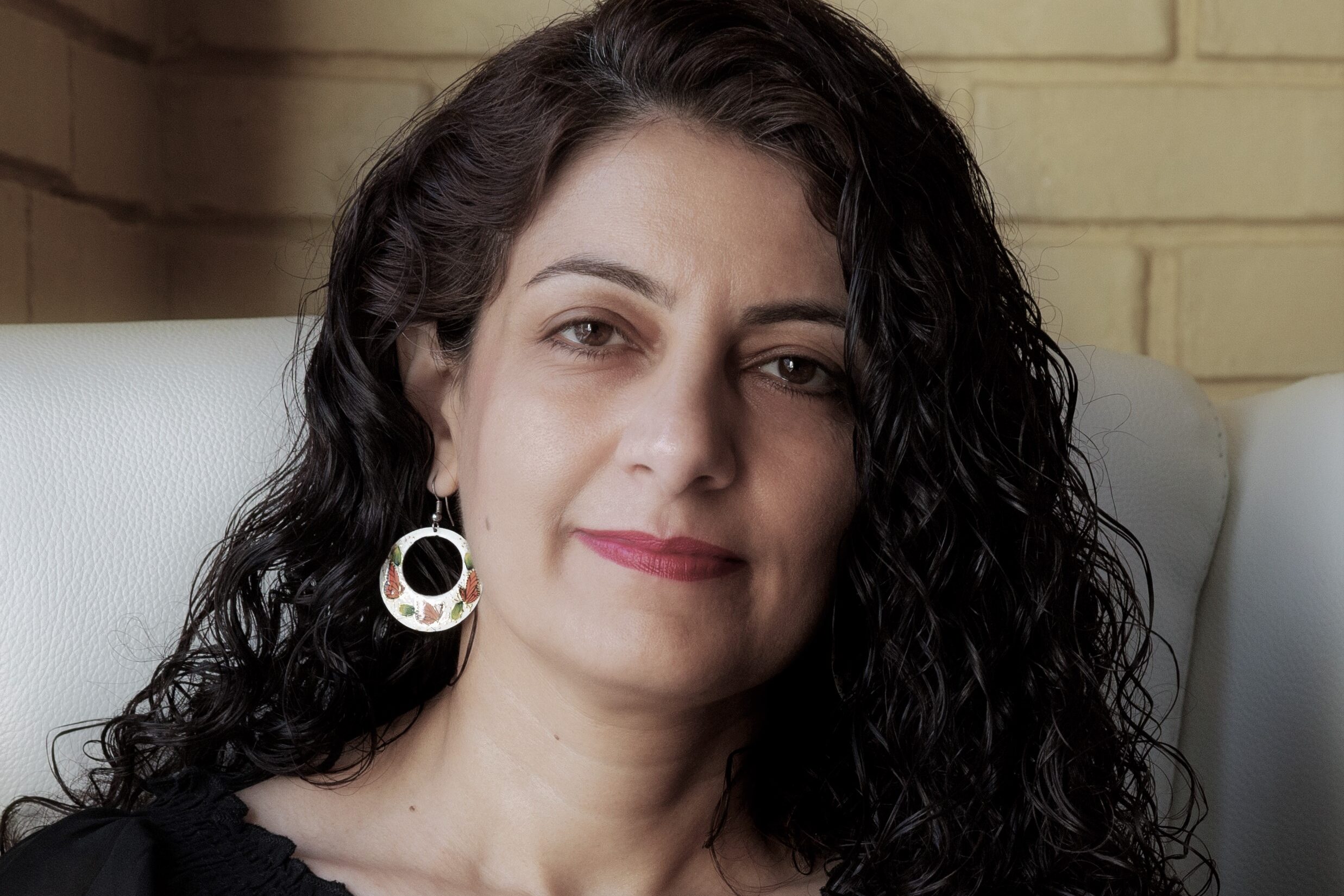 A woman with curly black hair and hoop earrings sits on a white chair. She is wearing a black top with embroidered details and has a slight smile. The background features a brick wall.