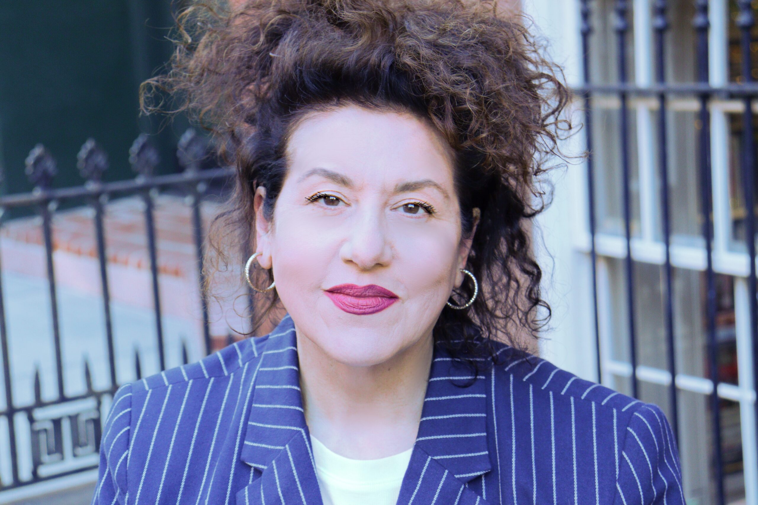 A person with curly hair and hoop earrings smiles slightly, wearing a pinstripe blazer and a white shirt. They are standing in front of a wrought iron fence.
