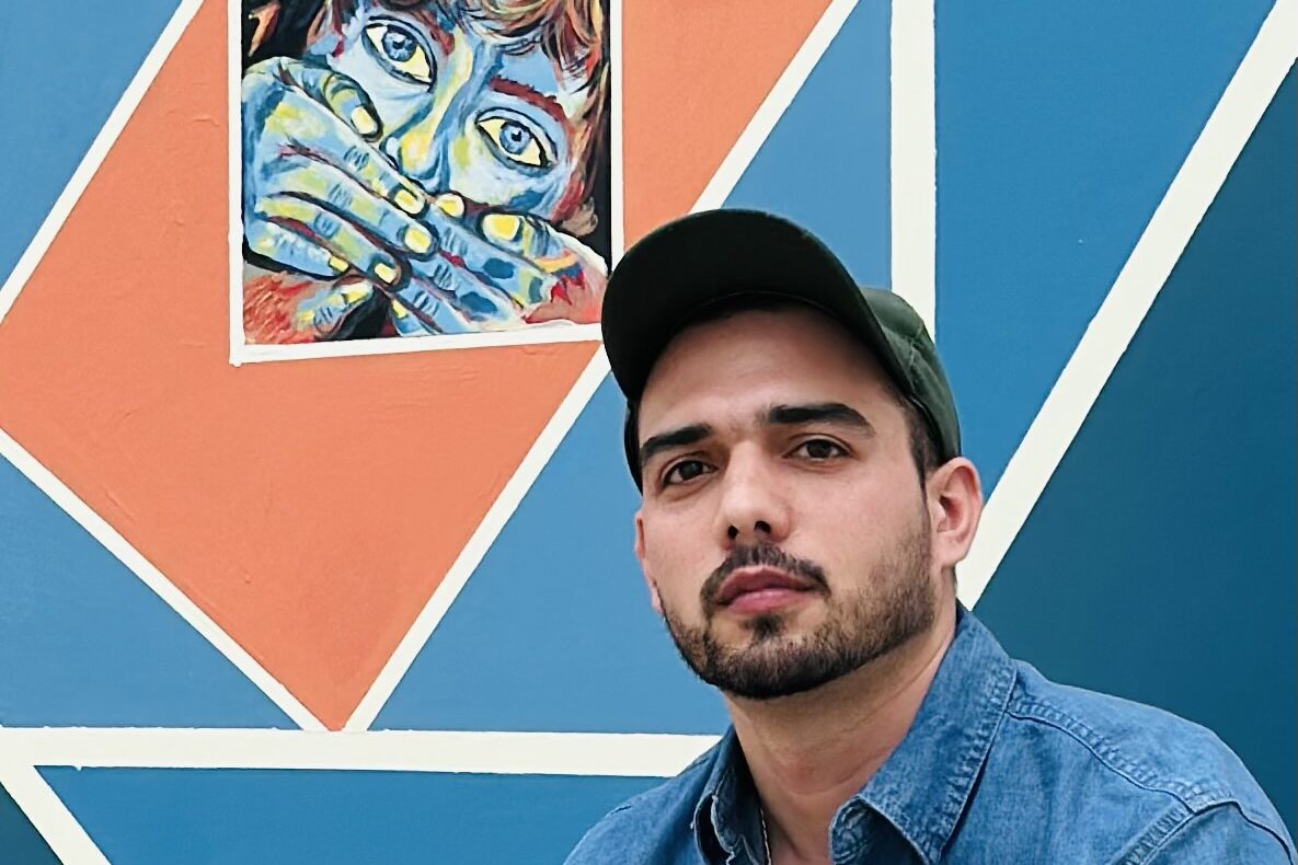 Man in a denim shirt and cap sitting in front of a colorful geometric mural featuring an expressive painting of a person covering their mouth.