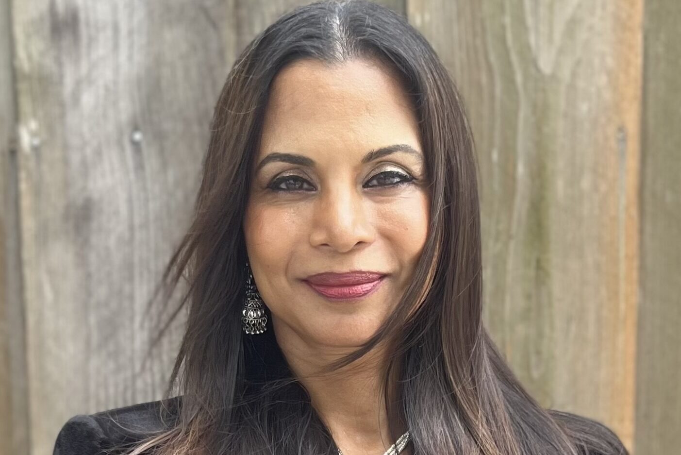 A woman with long, dark hair and a black jacket stands in front of a wooden wall. She is wearing earrings and a necklace, and she is smiling softly at the camera.