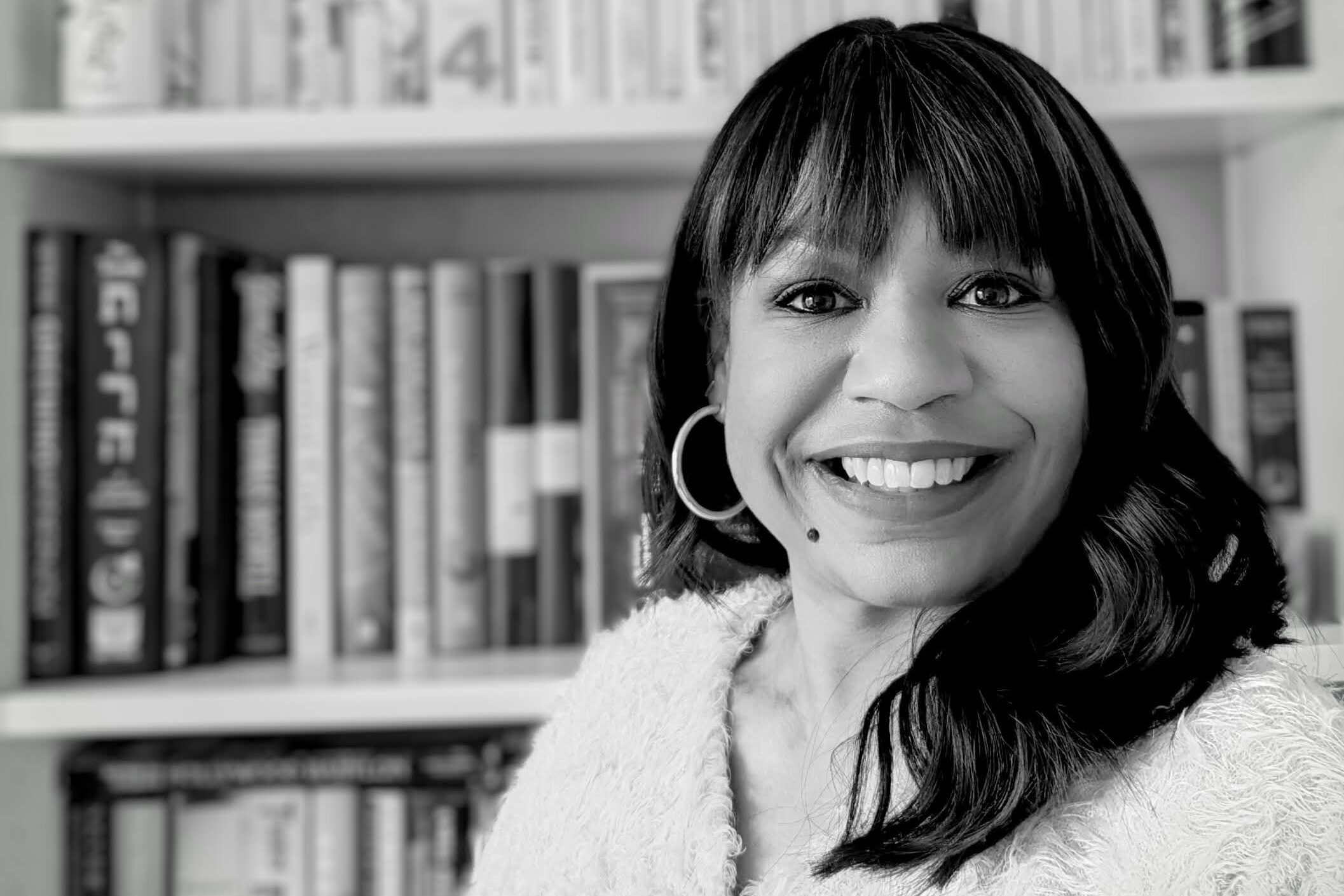 A woman with long dark hair, wearing a light-colored outfit, smiles at the camera. She stands in front of a bookshelf filled with various books. The image is in black and white.