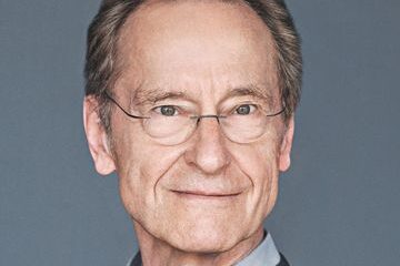 An older man with glasses smiles softly against a gray background. He is wearing a dark sweater over a light shirt.