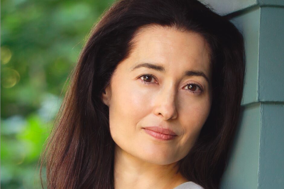A woman with long dark hair leans against a green wall, looking towards the camera with a slight smile. The background is blurred greenery, giving the image a peaceful, natural feel.