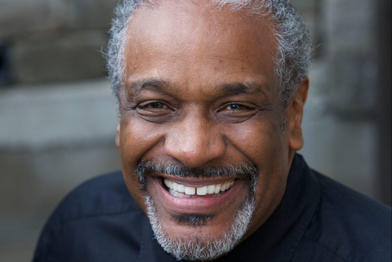Smiling man with gray hair and beard, wearing a black shirt and tie, standing in front of a blurred brick background.