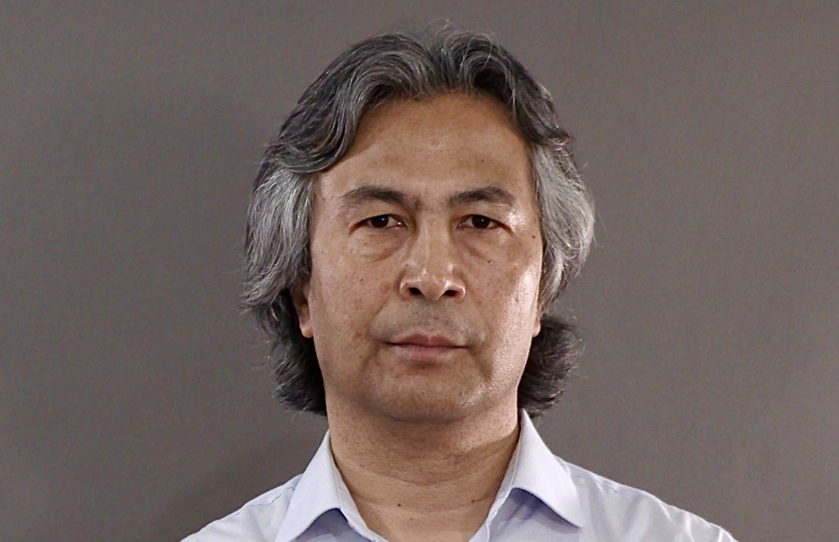 A person with shoulder-length gray hair is wearing a light blue collared shirt. They are looking directly at the camera. The background is a plain gray.