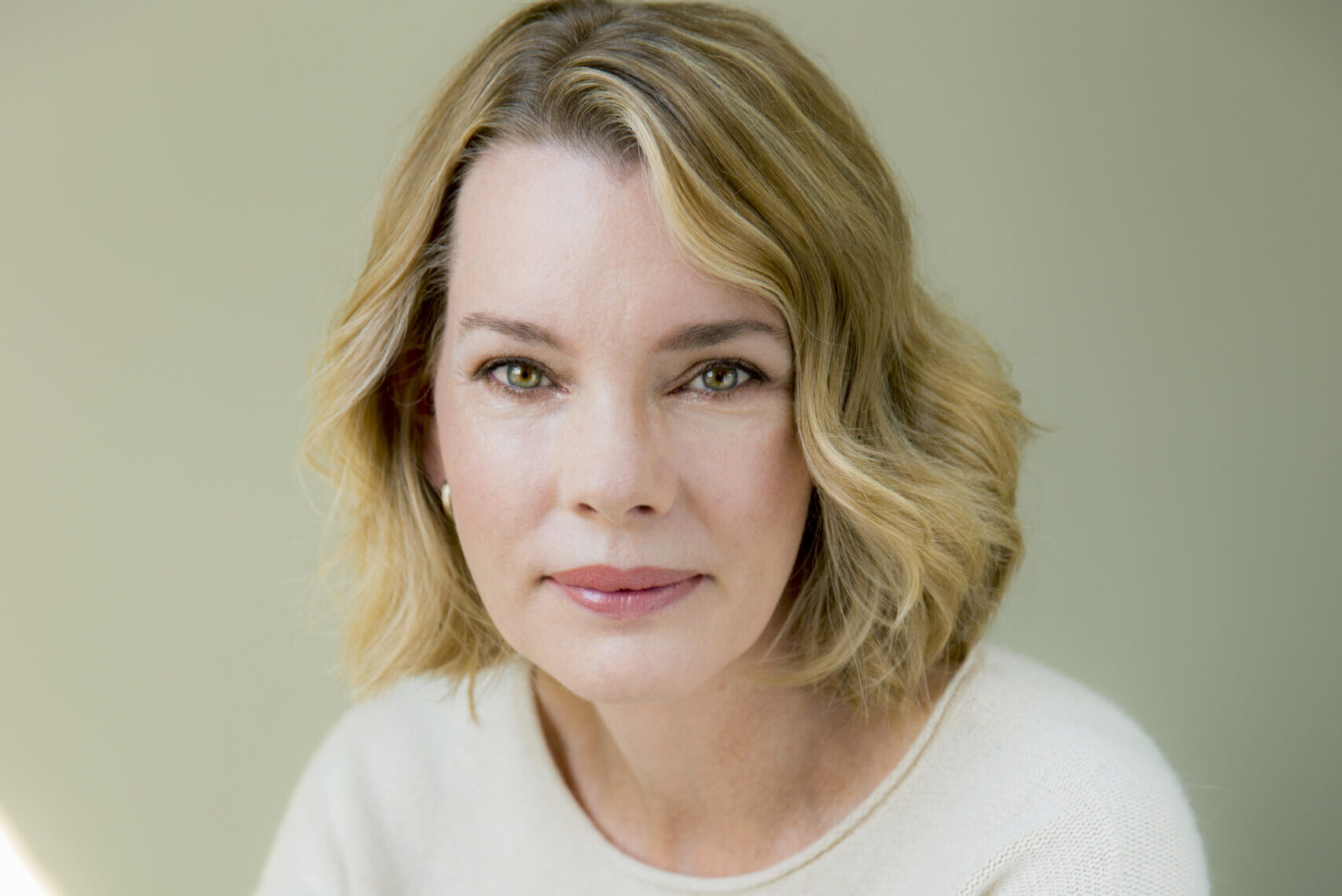 A woman with shoulder-length blonde hair sits against a light background, wearing a cream sweater and jeans. Her arms are crossed, and she has a calm, composed expression.