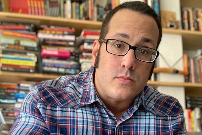 Person in a blue and red plaid shirt sits with hands together, looking at the camera in front of a full bookshelf.