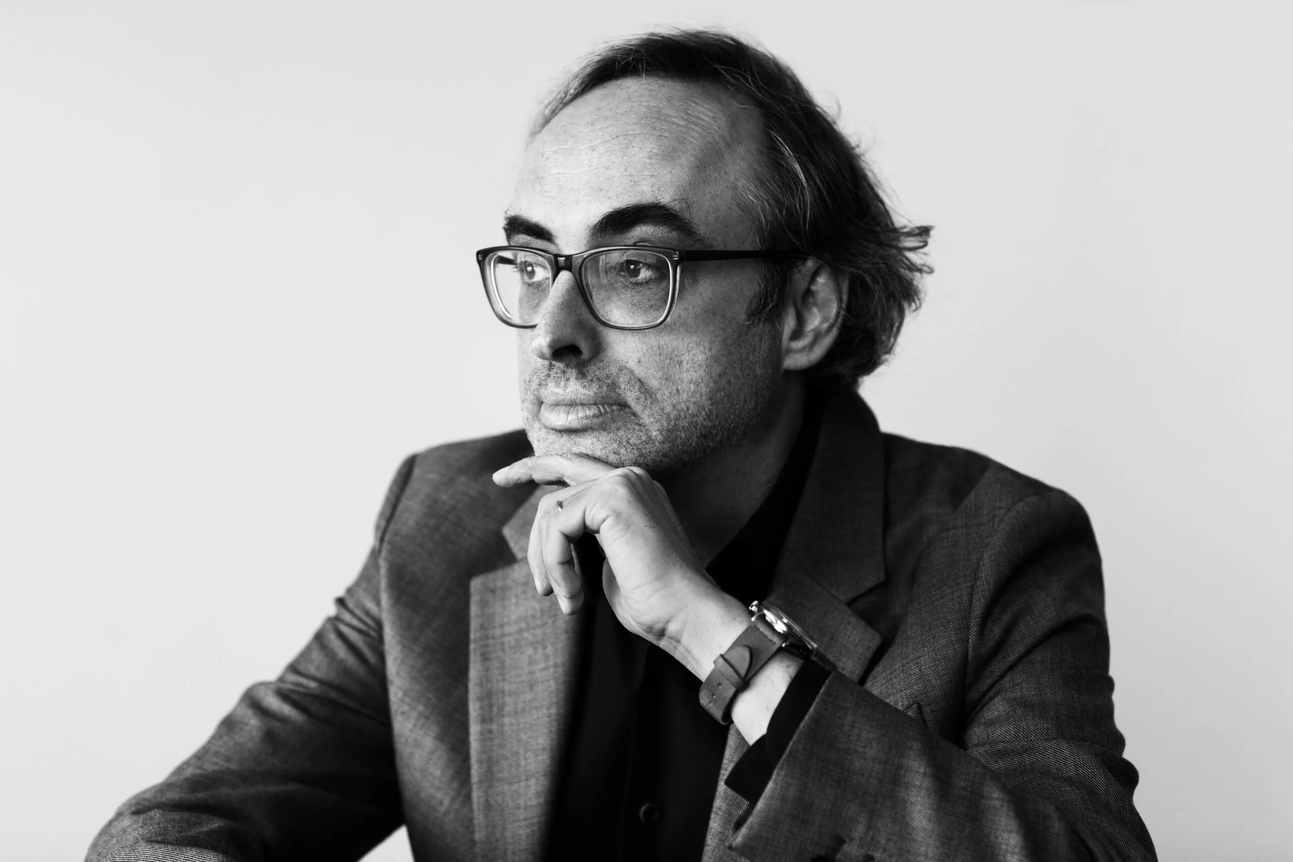 A man with glasses and a beard, wearing a suit, sits thoughtfully with his chin resting on his hand, looking to the side. The image is black and white with a plain background.