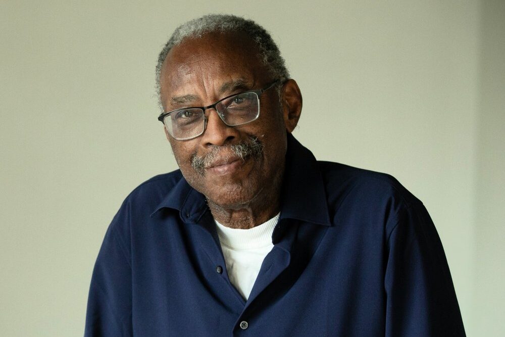 An elderly man with glasses and a mustache is wearing a dark blue shirt over a white t-shirt. He stands with a relaxed expression in a softly lit, neutral-colored room.