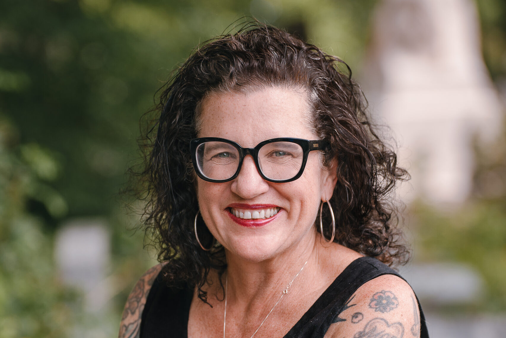 A woman with curly hair and black glasses smiles outdoors. She wears a black sleeveless top, showcasing her tattooed arms. She has hoop earrings, bracelets, and a cross necklace, standing with arms crossed in a lush, green setting.
