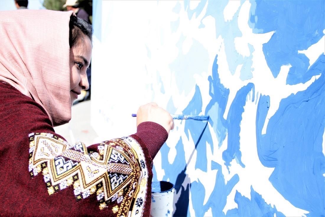 A person in a patterned sweater and headscarf is painting white bird silhouettes on a blue background. They hold a small brush, focusing intently on their work. A paint bucket sits nearby on the ground.