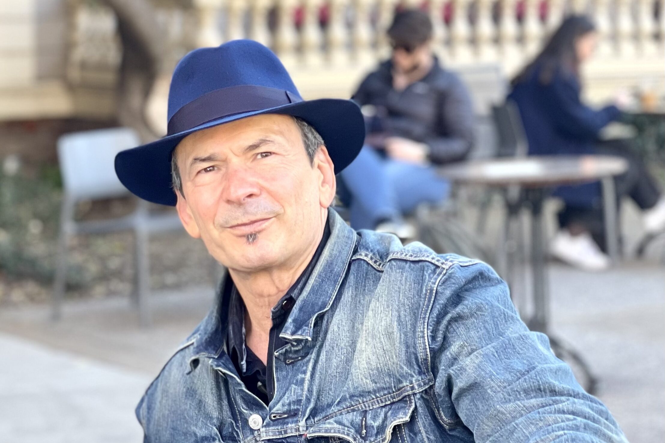 A person wearing a blue hat and denim jacket sitting outside. A porch with a railing is in the background, along with people sitting at tables, engaged in conversation or activities. The setting appears relaxed and casual.