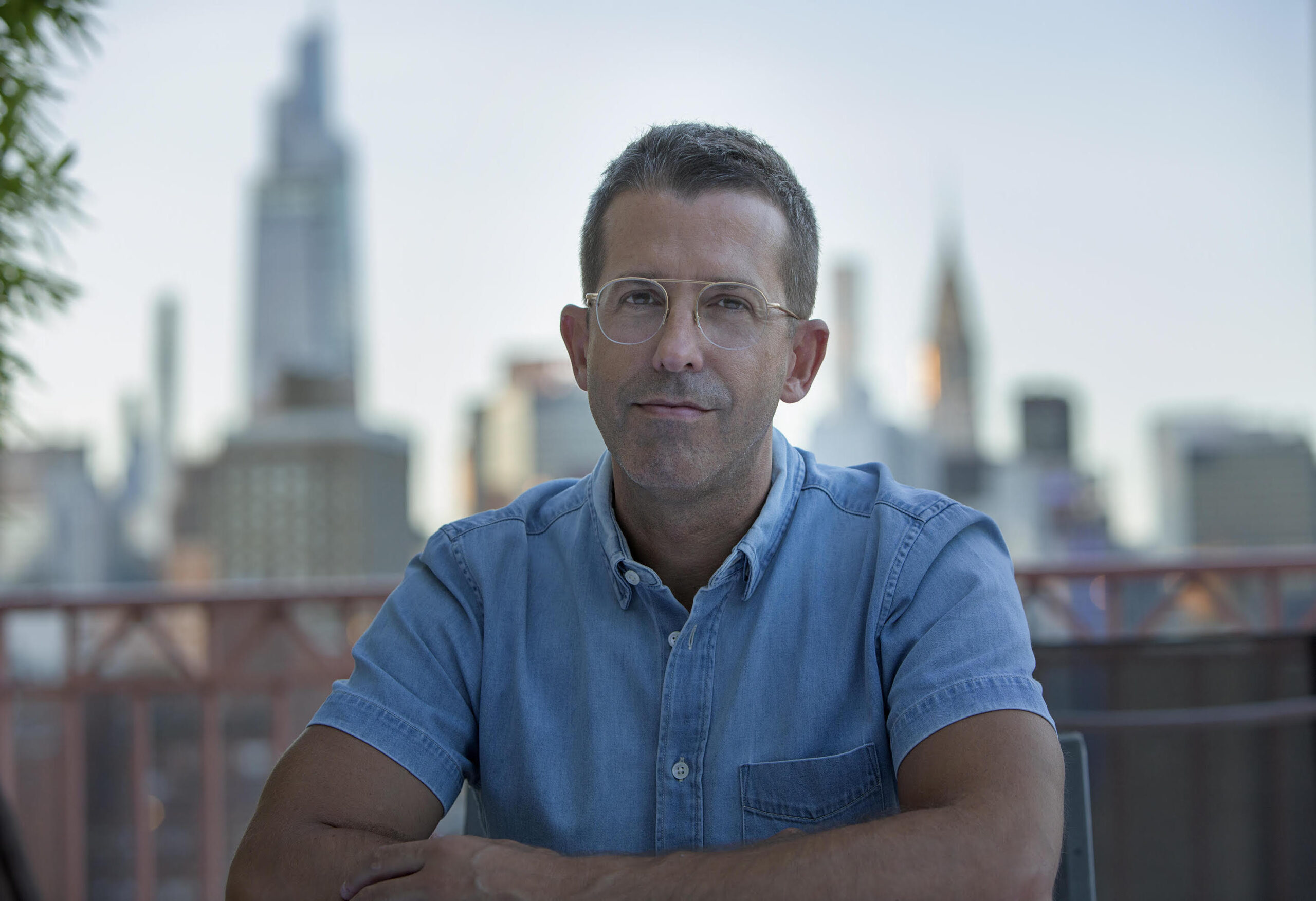 A man in glasses and a blue short-sleeve shirt sits at a table outdoors with a blurred city skyline in the background. The lighting is soft, suggesting a sunset or sunrise.