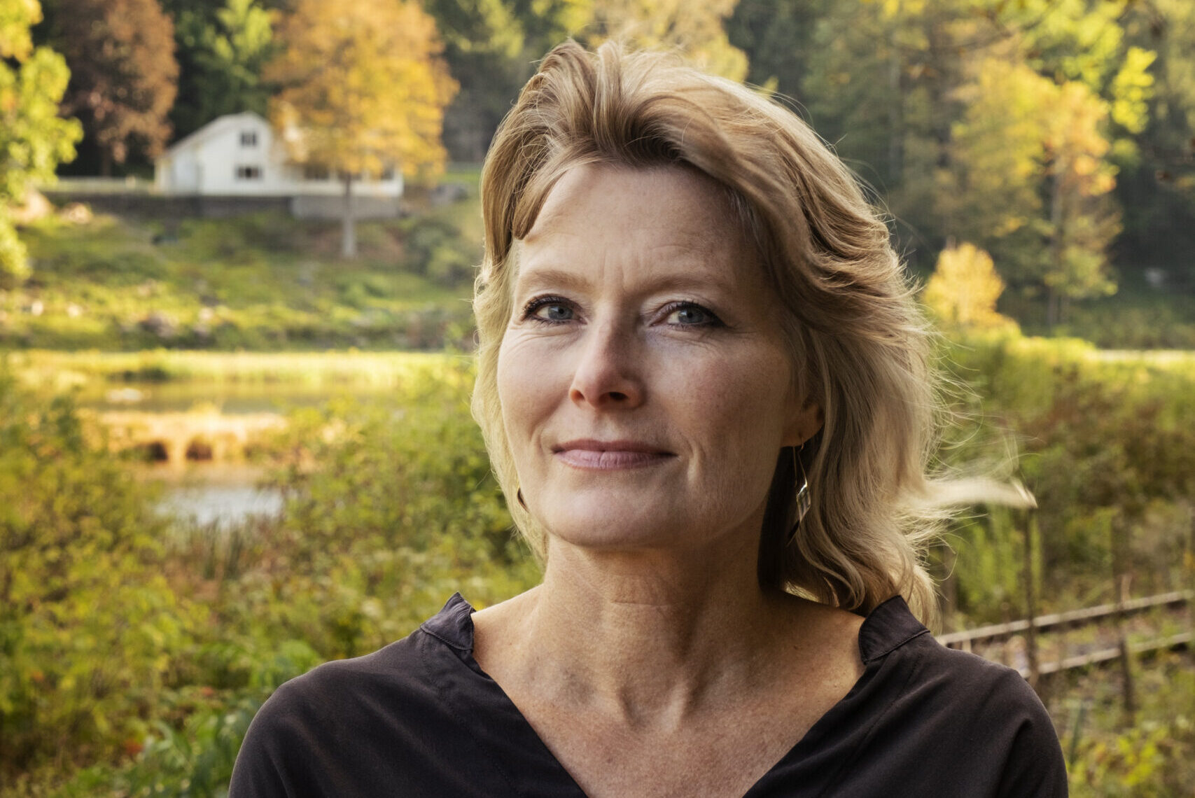 Woman in a black blouse stands outdoors with arms crossed. Behind her, a lush green landscape with autumn trees and a house in the distance. She gazes thoughtfully into the distance.