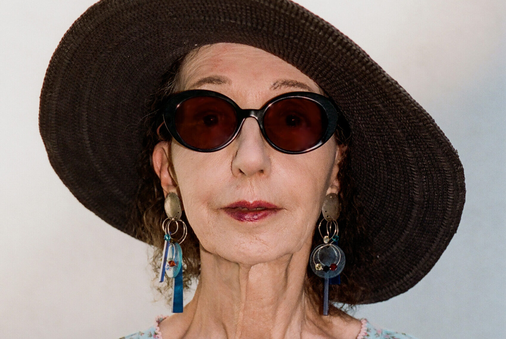 An older woman with wavy hair wears large brown sunglasses and a wide-brimmed hat. She dons a light blue floral blouse, multiple silver rings, and dangling earrings, standing against a light background.