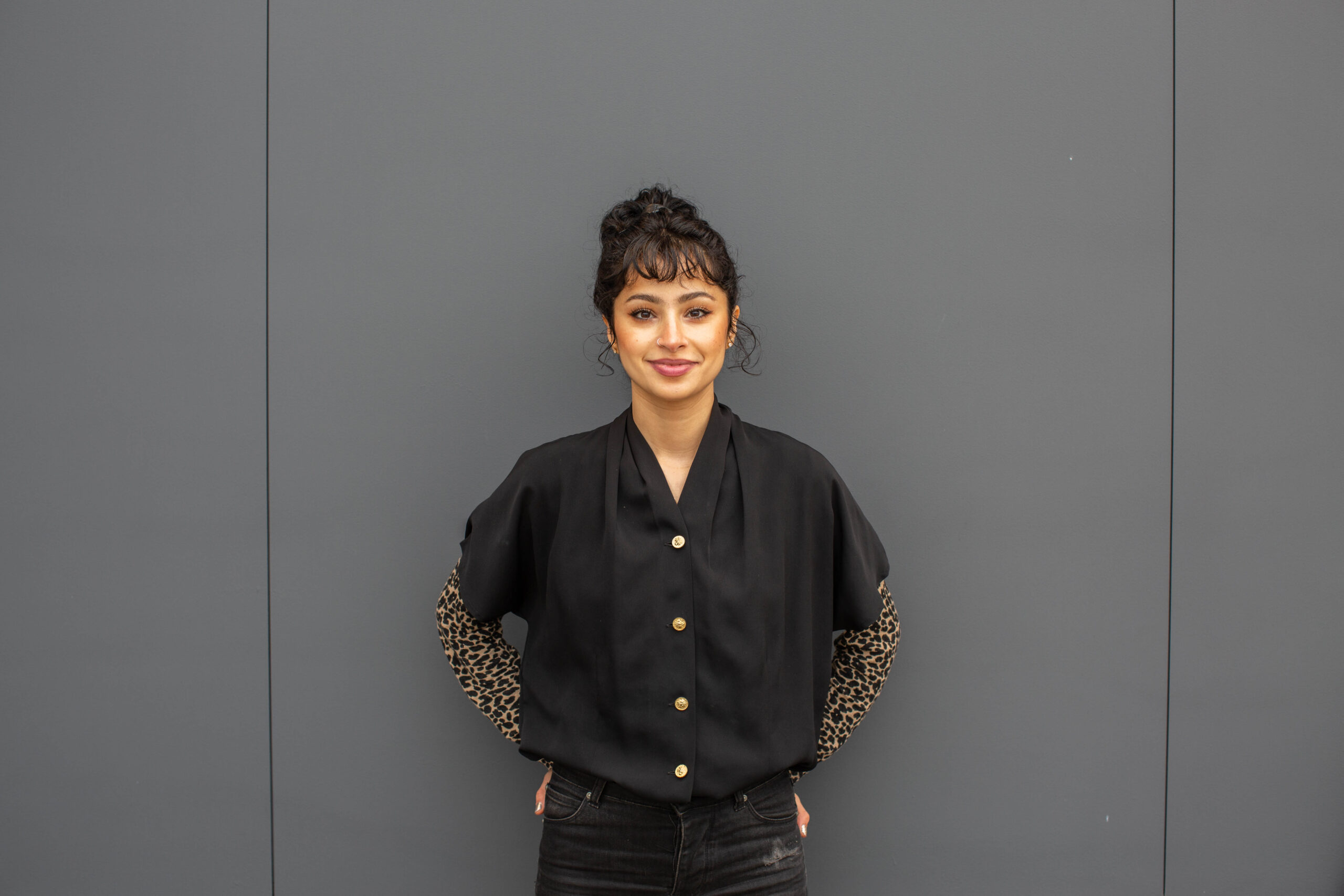 Woman with curly hair, wearing a black blouse with leopard print sleeves, stands against a plain gray background with hands in pockets, smiling gently.
