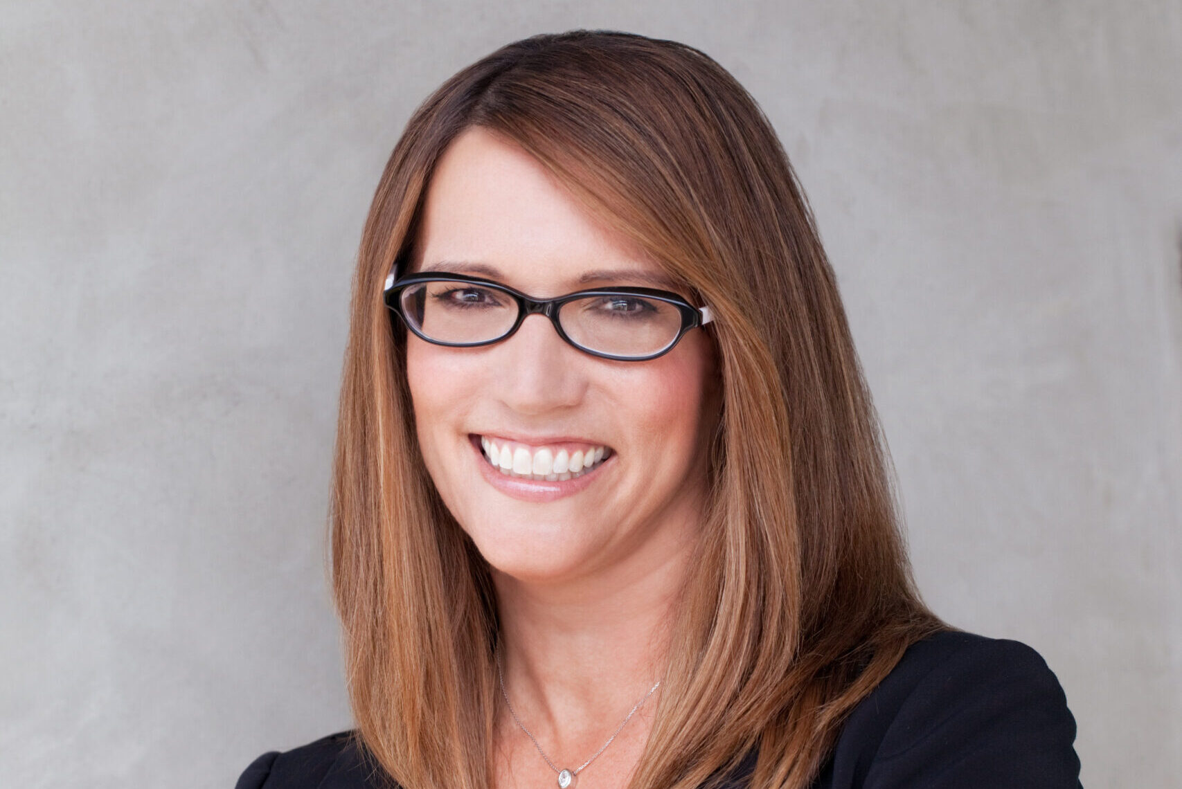A person with shoulder-length brown hair and glasses smiles at the camera. They are wearing a black blazer over a blue top, and their arms are crossed. The background is a plain, light-colored wall.