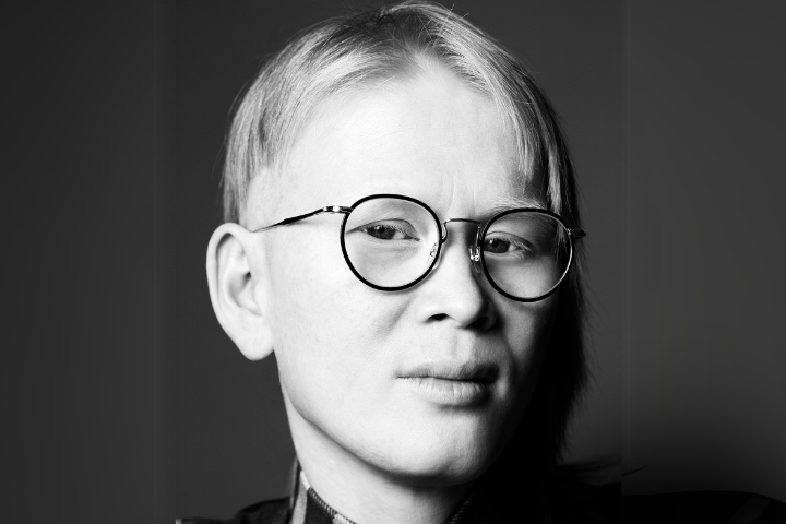 Close-up black and white portrait of a person with short, light hair and round glasses, looking into the camera with a neutral expression against a plain background.