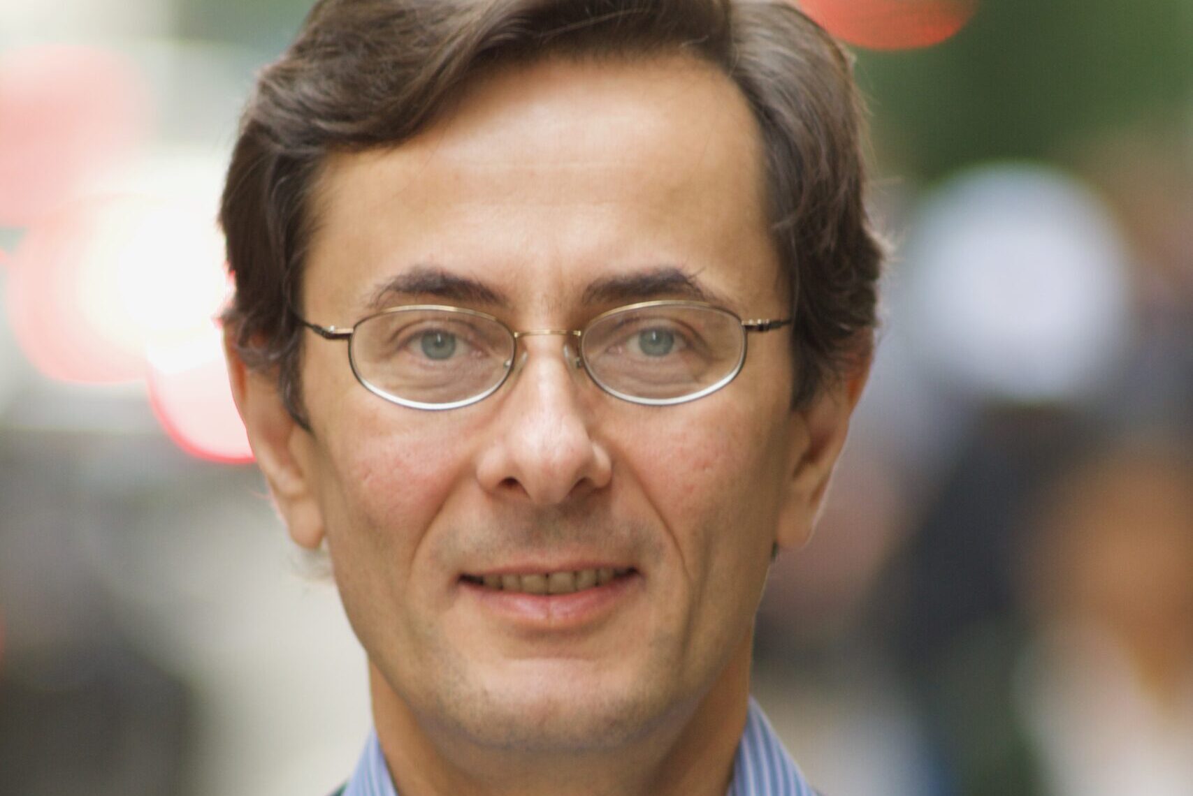 A person with glasses, short brown hair, and a faint smile is wearing a blue striped shirt and a dark blazer. The background is blurred, showing some hints of city life with lights and indistinct shapes of people.