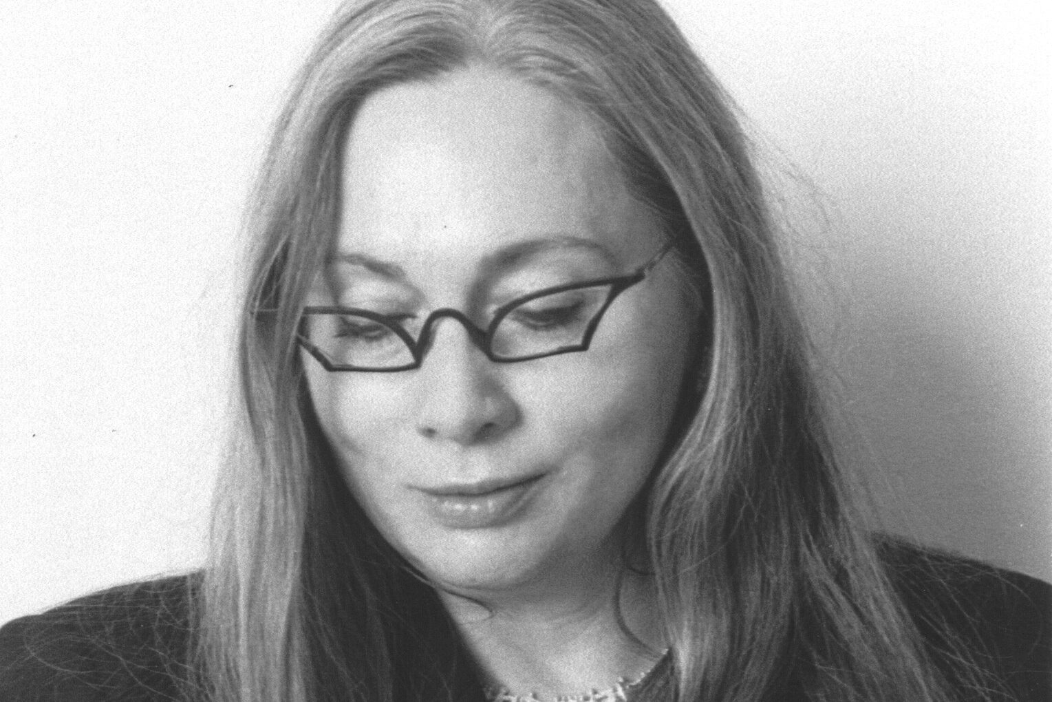 A black and white photo of a person with long hair wearing glasses, a blazer, and various jewelry, including rings and bracelets. They look down, with one hand on their hair. The background is plain.