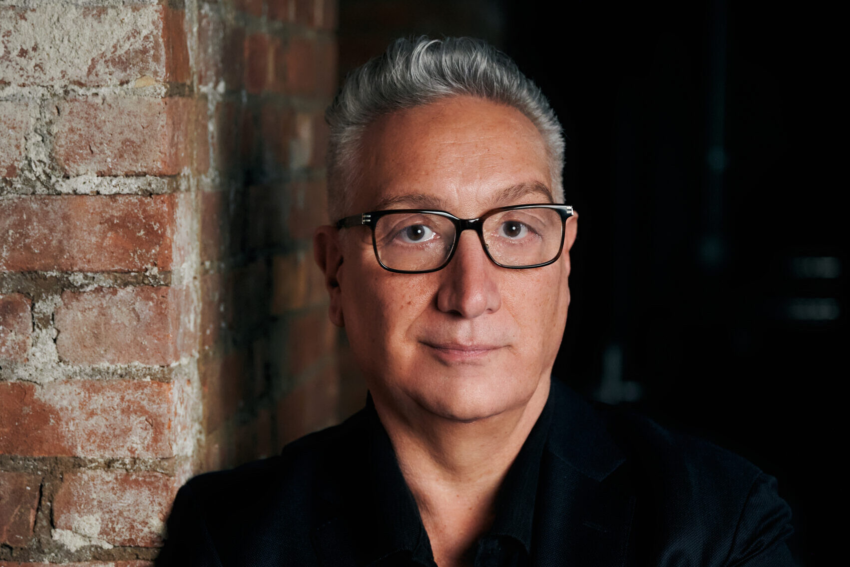 A person with short gray hair and glasses stands against a brick wall, arms crossed. Theyre wearing a black suit and have a calm expression, set against a dimly lit background.