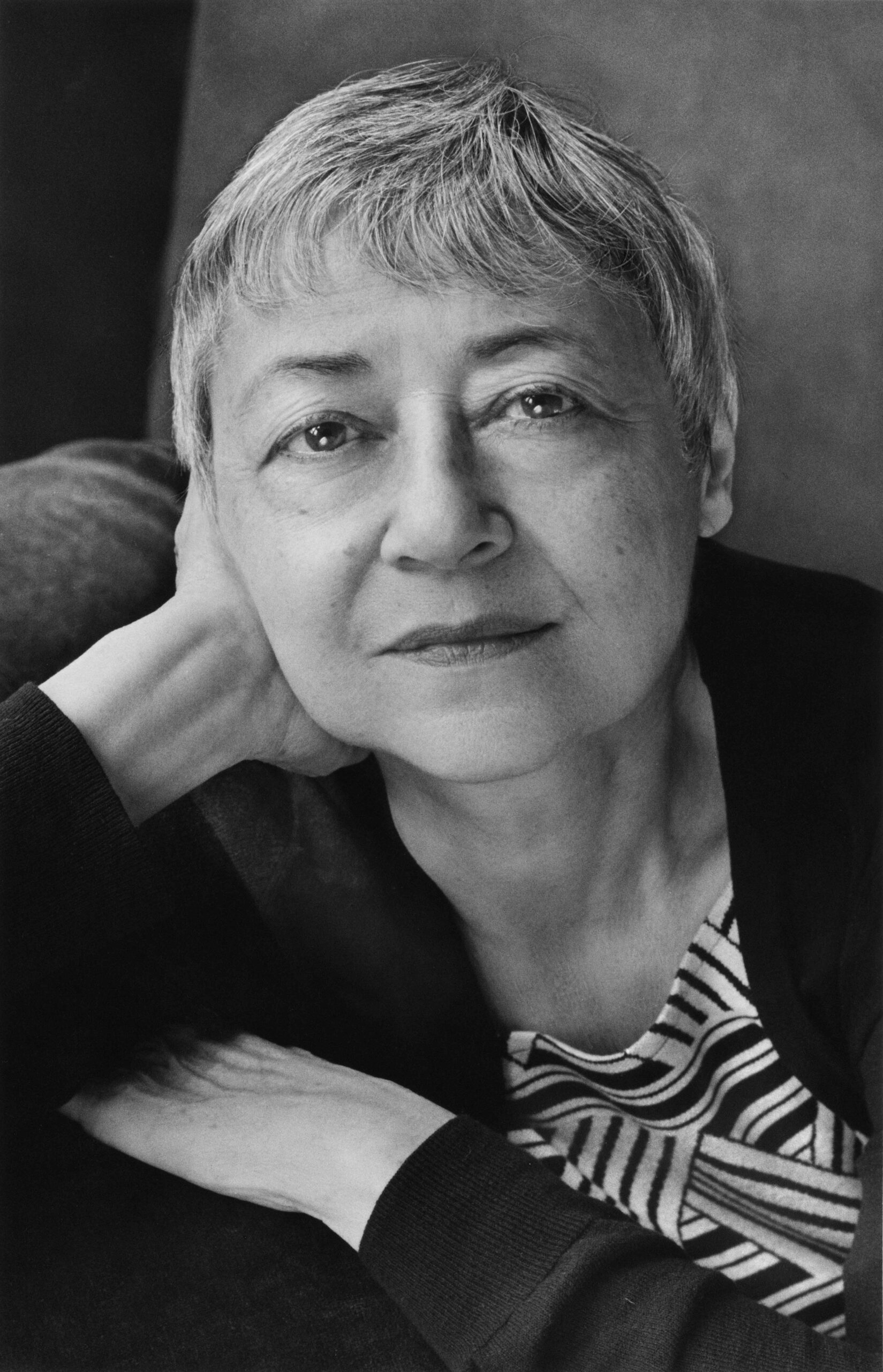 Black and white portrait of a person with short hair, resting their head on their hand. They wear a patterned top and have a serene expression, gazing directly into the camera.