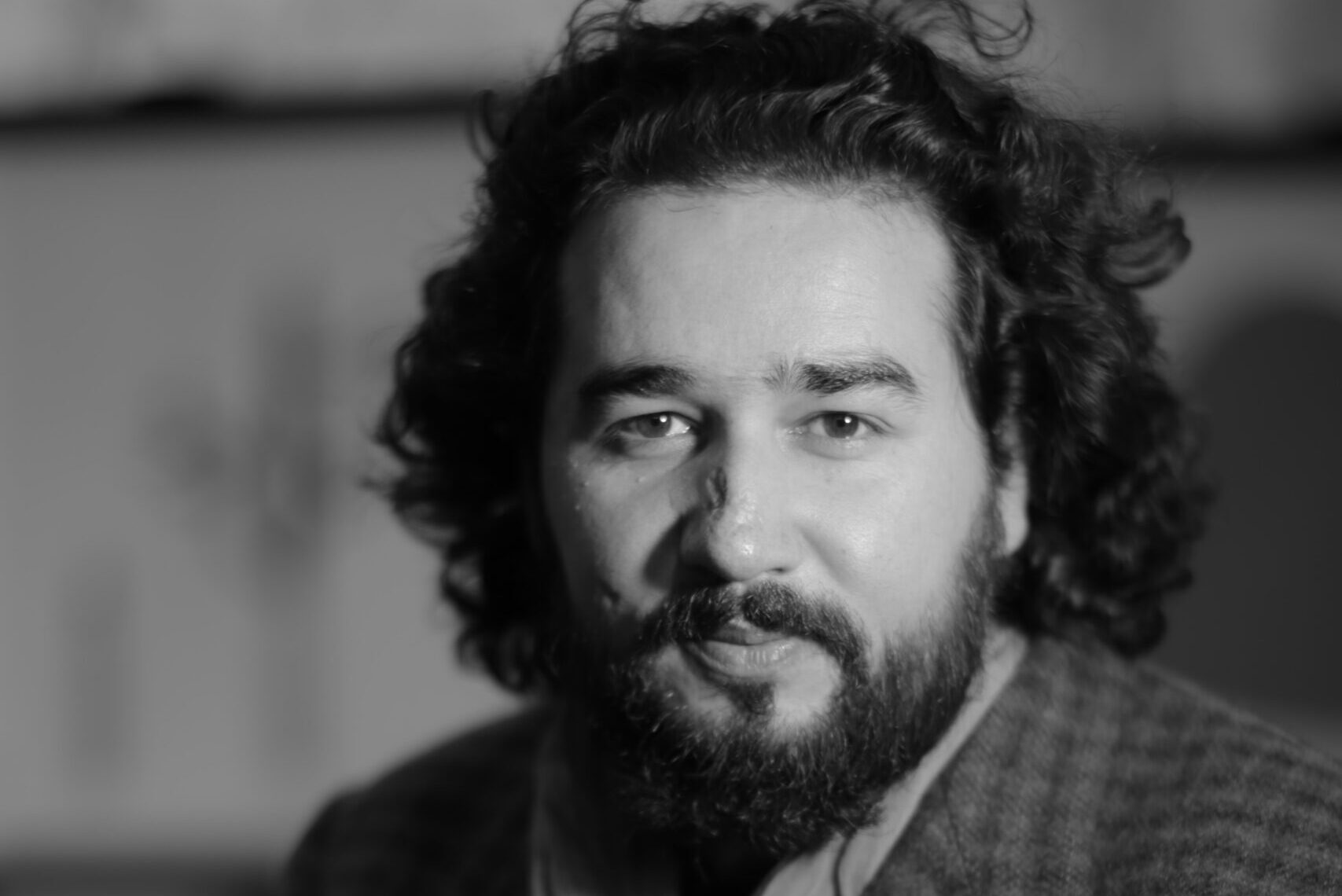 A black-and-white portrait of a bearded man with curly hair, wearing a patterned blazer, a shirt, and a scarf. He looks at the camera with a calm expression, set against a blurred indoor background.