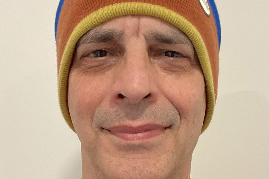 A person wearing a colorful beanie with brown, orange, and blue stripes. Theyre dressed in a dark sweater and a gray shirt, standing against a plain light-colored background.