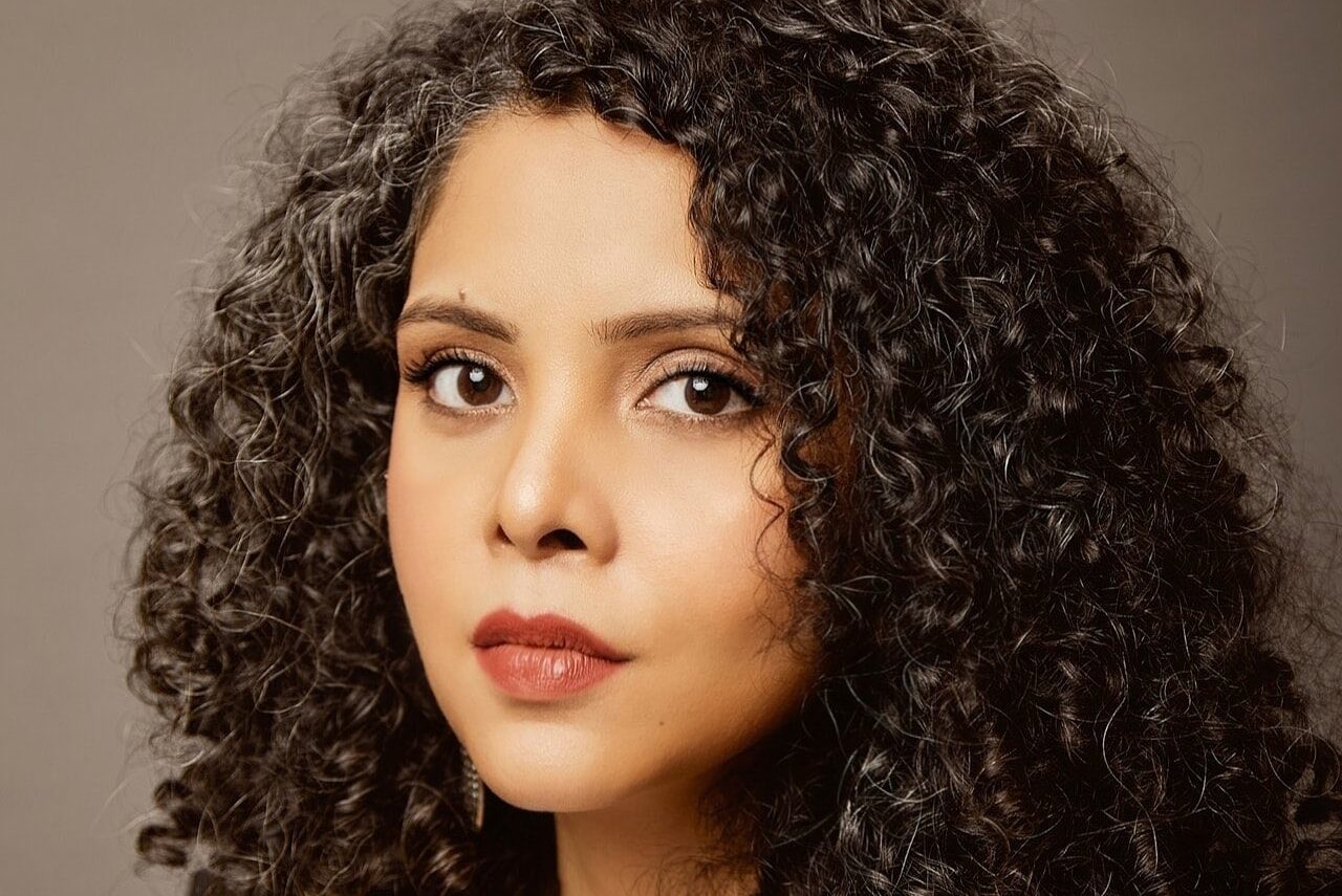 A person with curly hair, wearing a white top and dark blazer, gazes confidently at the camera against a neutral background.