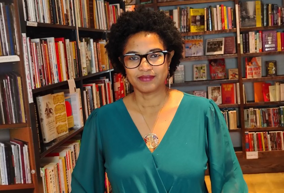 A person with curly hair, wearing glasses and a green top, stands in a library surrounded by bookshelves filled with various books.