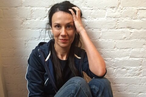 A woman with long dark hair sits against a white brick wall, wearing a dark jacket, jeans, and red socks. She rests her head on her hand and gazes at the camera, sitting on a wooden floor.