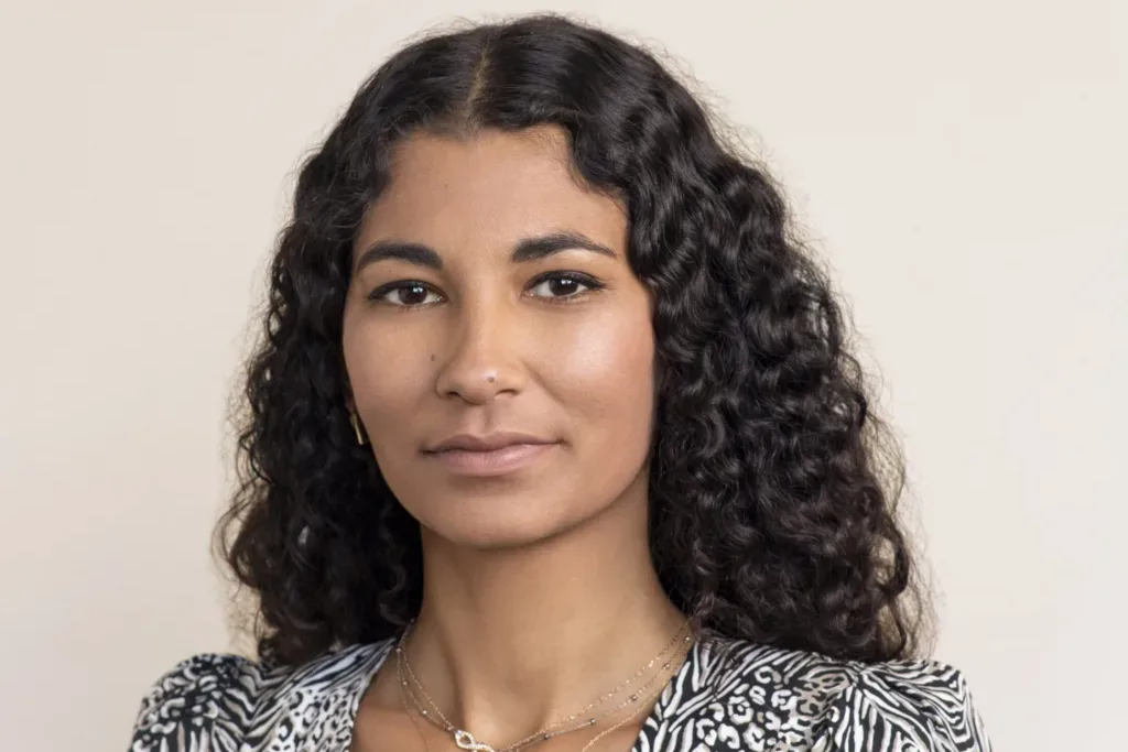 Young person with long, curly hair and a neutral expression, wearing a patterned blouse and layered necklaces, against a plain background.