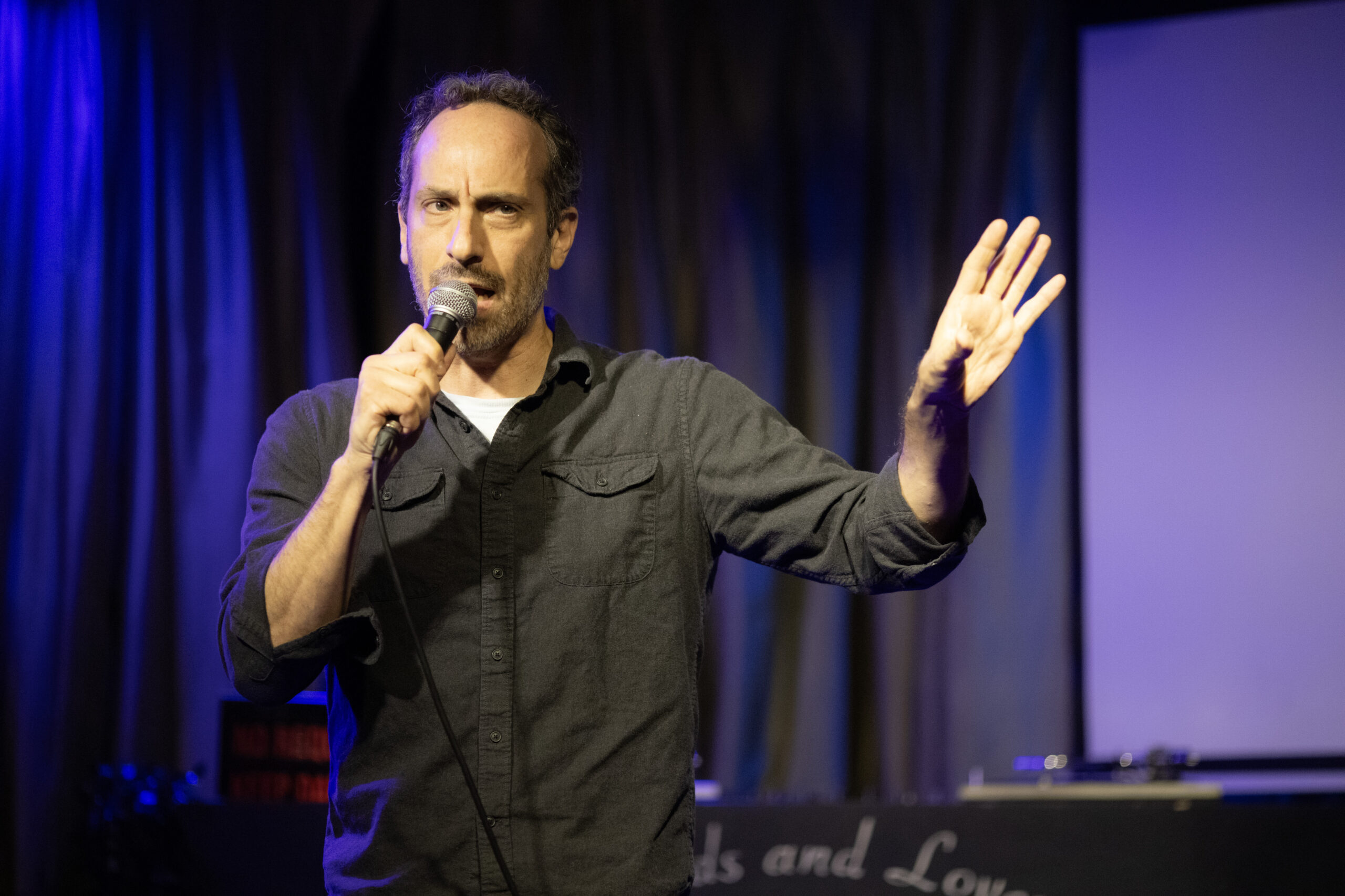 A man holding a microphone stands on stage under blue and yellow lights, speaking with one hand raised, in front of a curtain and a large blank screen.