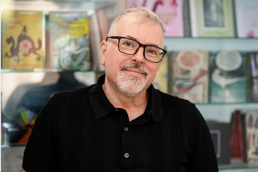 A man with short gray hair and glasses is wearing a black polo shirt. He is standing in front of a bookshelf filled with various books and decorative items, looking at the camera with a slight smile.