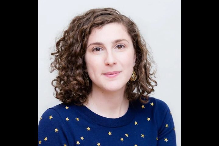 A woman with curly brown hair, wearing a navy blue sweater with yellow stars and gold earrings, sits against a light background, looking at the camera with a soft smile.