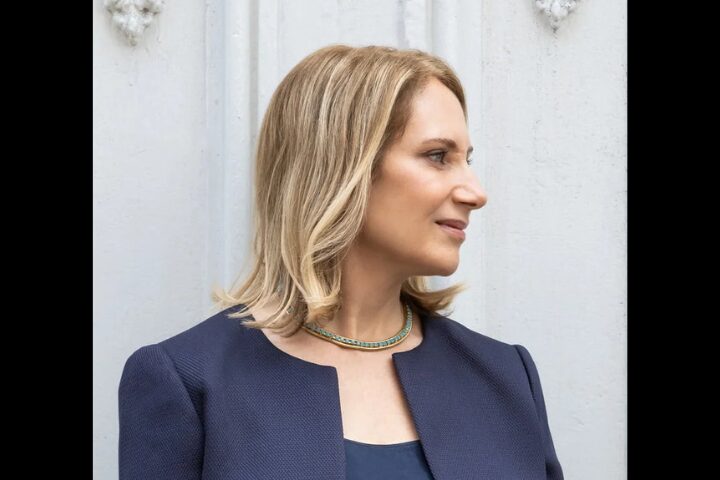 Woman with shoulder-length blonde hair wearing a blue jacket and green necklace, looking to the right, standing in front of a white textured wall.