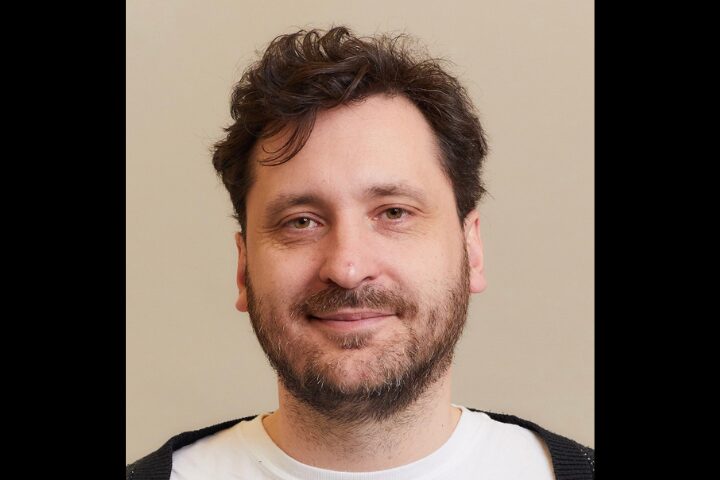 A bearded man with wavy, dark hair is smiling gently at the camera. He is wearing a white shirt and a black cardigan. The background is a plain, beige color.
