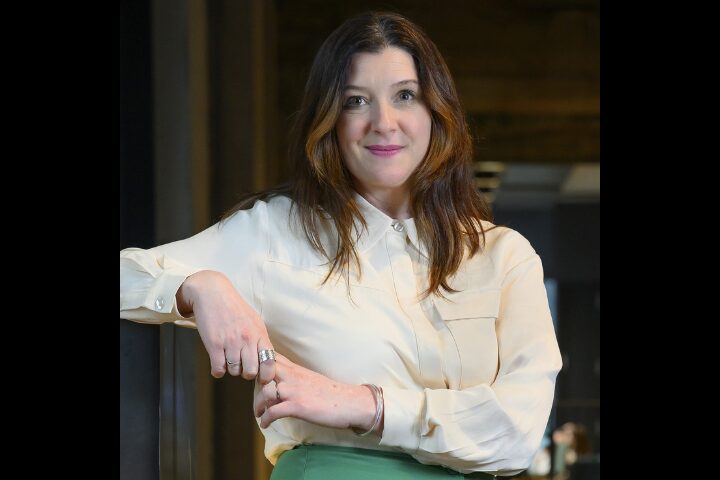 A confident woman with long brown hair stands indoors, leaning against a surface. She is wearing a cream-colored button-up shirt and a green skirt. The background is dimly lit, suggesting an office or professional setting.