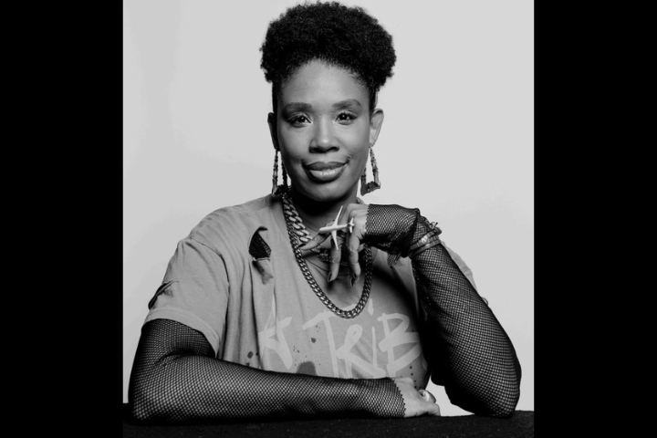A woman with short curly hair sits at a table, resting her arm on it. She wears a t-shirt, fishnet gloves, chunky jewelry, and earrings. She is looking confidently at the camera against a plain background.