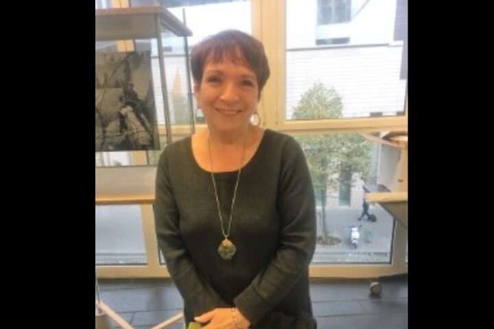 A woman with short brown hair, wearing a dark sweater and a long necklace, smiles while standing indoors in front of large windows with a street view outside.
