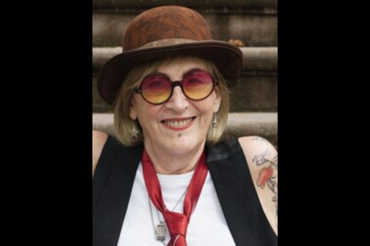 An older person with blond hair smiles, wearing a brown hat, round pink and yellow glasses, a red tie, white shirt, black vest, and showing a tattooed arm. Stairs are visible in the background.