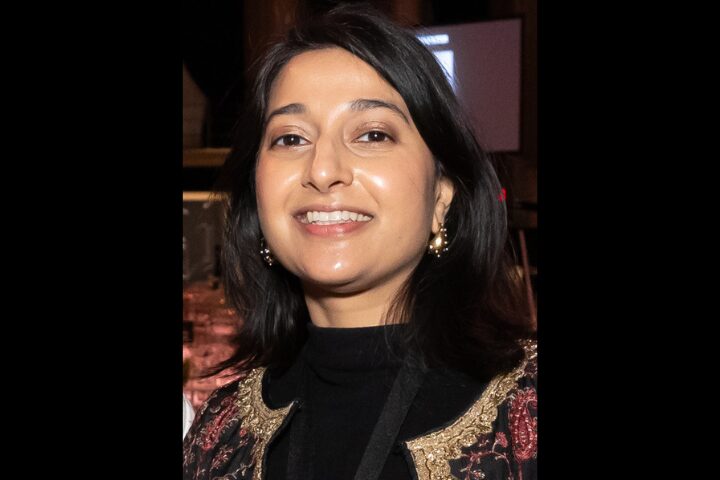 A woman with dark hair, wearing a black top and an ornate jacket, smiles at the camera. She is indoors, with a blurred background featuring dark and light tones.