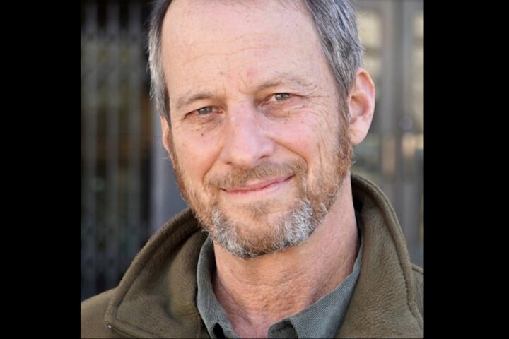 A middle-aged man with gray hair and a beard is smiling gently. He is wearing a green jacket over a collared shirt. The background is blurry and appears to be outdoors.